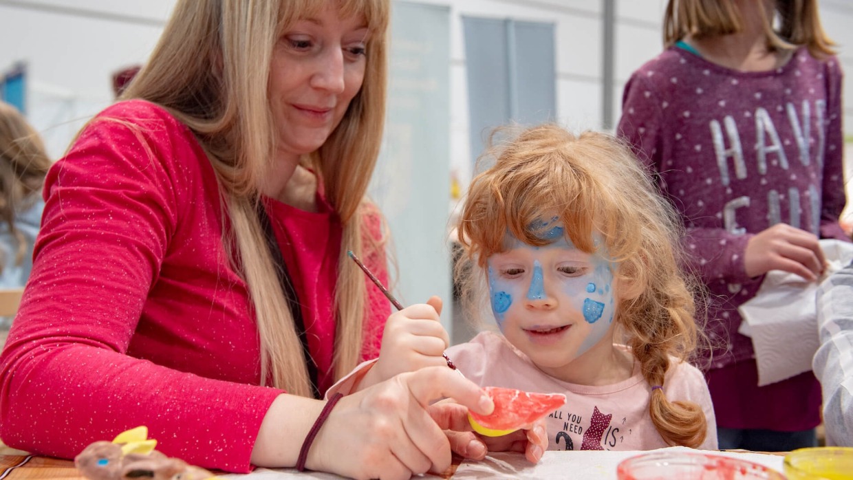 A woman is crafting with a small, face-painted child at a table.
