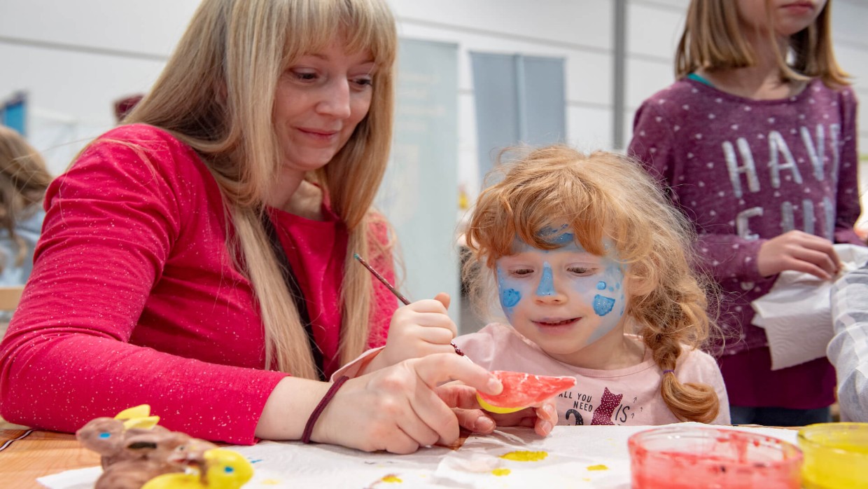 Eine Frau bastelt mit einem kleinen, bunt geschminkten Kind an einem Tisch.
