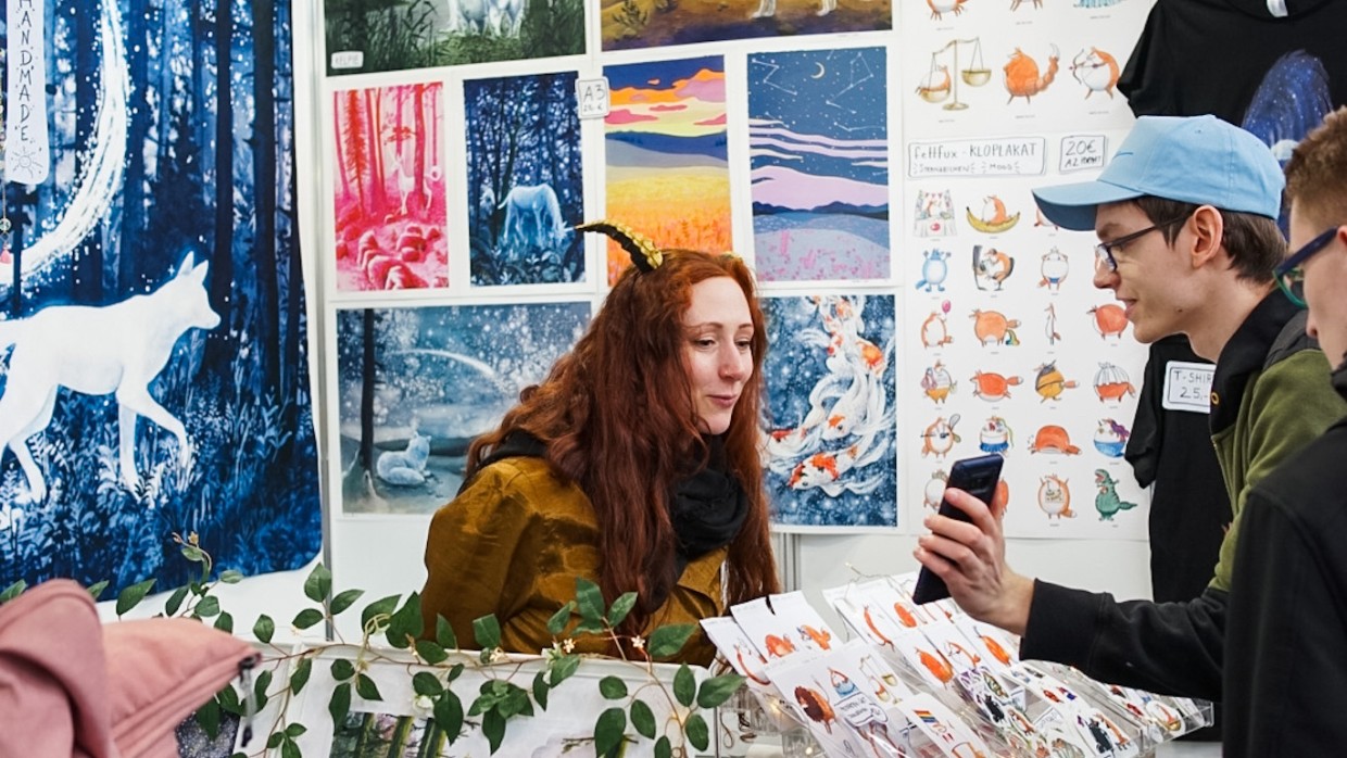 Besucher zeigt einer Ausstellerin am Messestand etwas auf dem Smartphone in der New Artist Alley auf der Manga-Comic-Con 2024