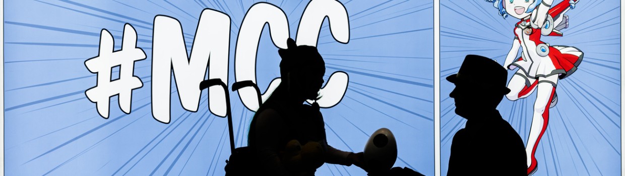 Die Silhouetten von 2 Personen sind erkennbar vor einer leuchtenden Wand, die bedruckt ist im MCC-Stil mit Macoco. Eine Person sitzt im Rollstuhl, die andere kniet davor und überreicht ihr einen eiförmigen Gegenstand.