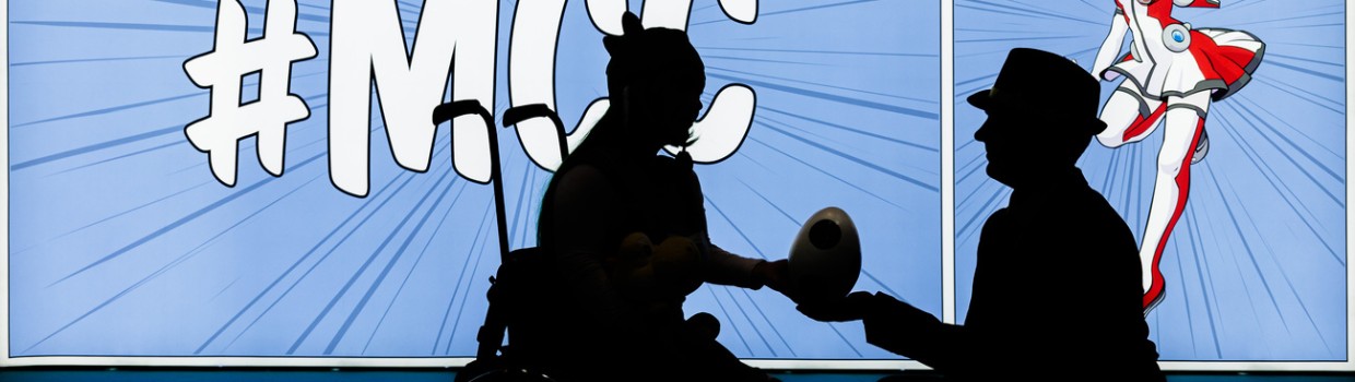 Die Silhouetten von 2 Personen sind erkennbar vor einer leuchtenden Wand, die bedruckt ist im MCC-Stil mit Macoco. Eine Person sitzt im Rollstuhl, die andere kniet davor und überreicht ihr einen eiförmigen Gegenstand.