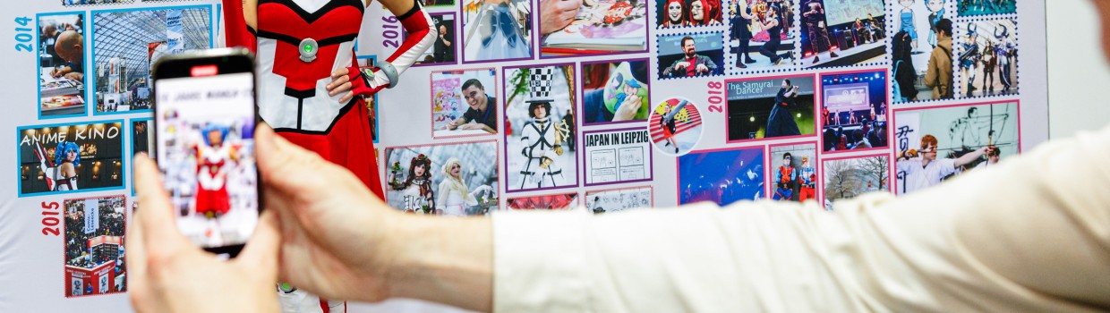 MaCoCo posiert vor einer Wand in der Jubiläumsausstellung der Manga-Comic-Con 2024 für einen Fotografen mit einem Smartphone