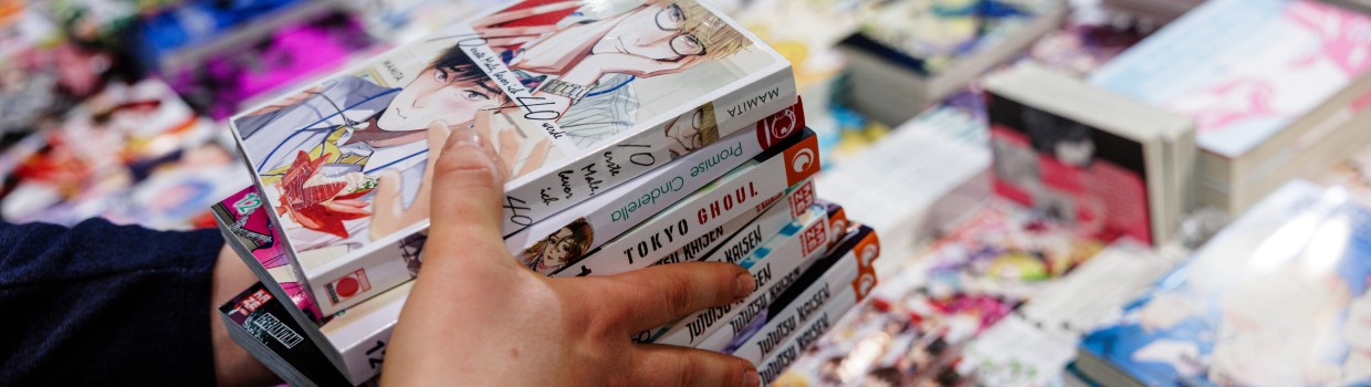 Hände halten einen Stapel Mangehefte über einem Tisch gefüllt mit einer Vielzahl an Manga auf der Manga-Comic-Con 2024