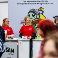 mehrere Personen vom Awareness Team SAM der Manga Comic Con 2025 stehen am Stand, Besucher:innen laufen daran vorbei
