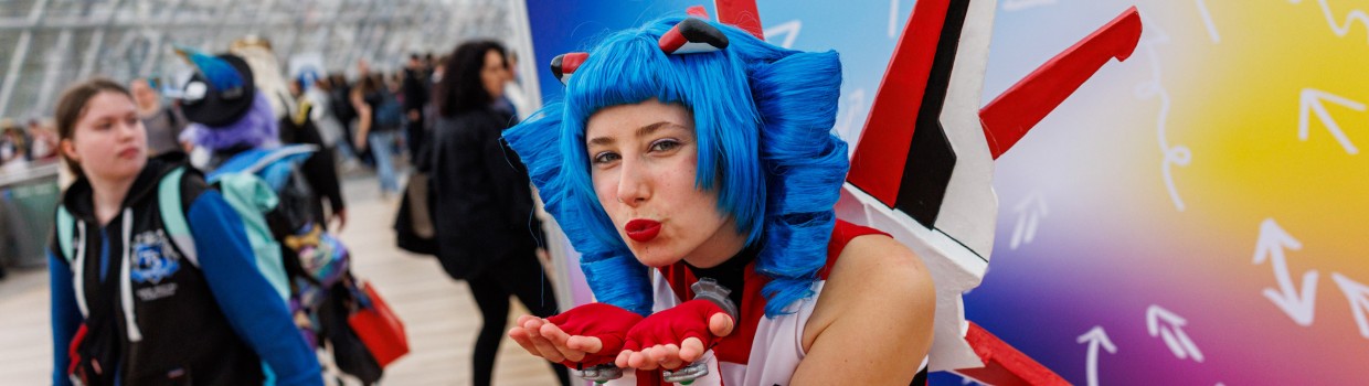 Das Maskottchen der Manga Comic Con Macoco macht einem Luftkuss vor einer bunten Leinwand auf der Manga Comic Con 2025