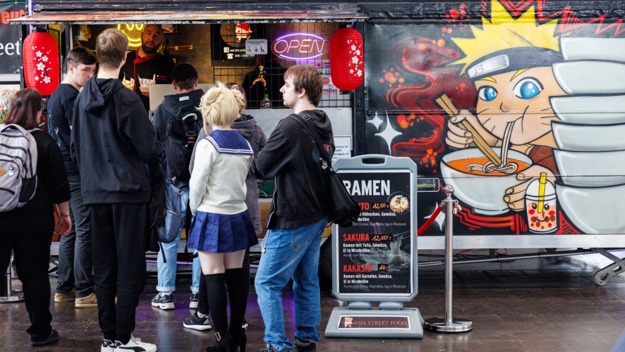 Several people are standing in line in front of a food truck at the Manga Comic Con 2025. The food truck is labeled Naruto cooks and features an illustration of a manga character eating noodles.
