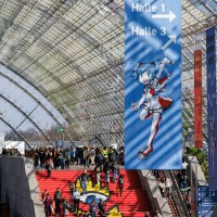 Crowds on the red staircase under a banner for Manga-Comic-Con 2025 with signposts and manga characters in the glass hall of the Leipzig Trade Fair.