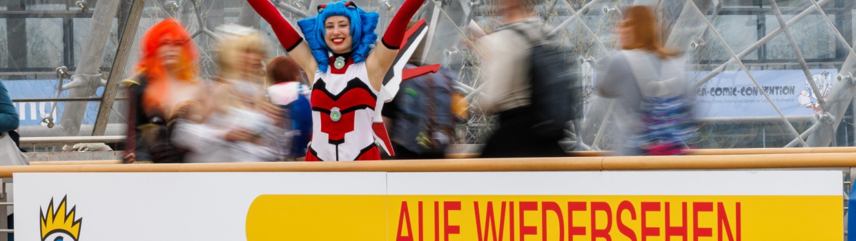MaCoCo poses on the gallery in the glass hall of the Leipzig Trade Fair in front of a banner displaying the date of Manga Comic Con 2026.