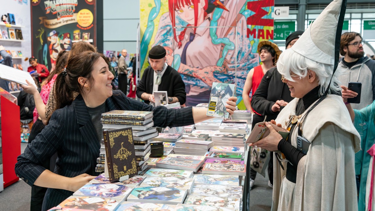 Ausstellerin überreicht Cosplayer:in ein Mangaheft über einen Verkaufstischer voller Manga auf der Manga Comic Con 2025