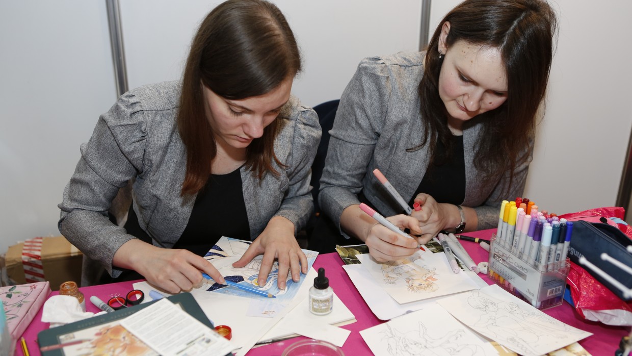 Zwei junge Frauen sitzen an einem Tisch und zeichnen. Auf dem Tisch liegen Papierbögen und Stifte, neben anderem Zeichenmaterial.